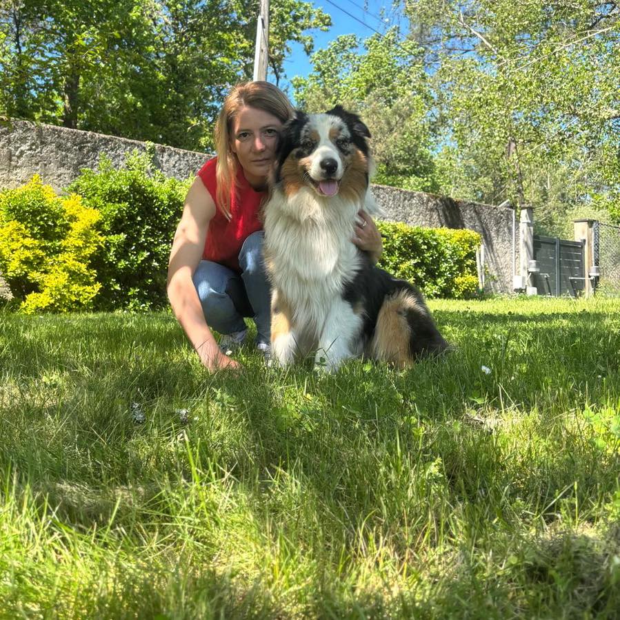 Femme accroupie dans l'herbe, tenant un Berger Australien au poil long et multicolore, dans un jardin ensoleillé.